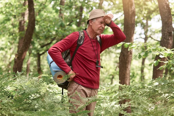 老年男子在野外森林里艰难、艰难、劳累、精疲力竭的探险，头疼难忍，手挽着额头，随随便便穿着，有睡垫和背包。 旅游概念. — 图库照片