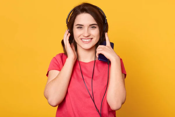 Tutup potret wanita muda yang cantik mendengarkan musik melalui headphone dan ponsel, melihat langsung ke kamera dan tersenyum terisolasi di atas latar belakang kuning, gadis tampak bahagia dan puas . — Stok Foto