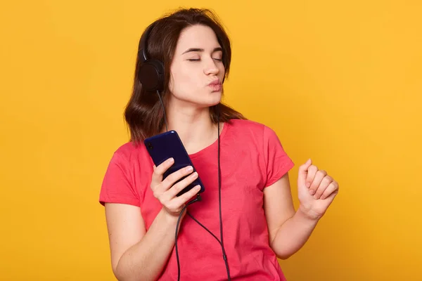 Fotografie orizontală a unei fete atractive cu păr întunecat, cu distracție izoaltată peste fundal galben în studio, doamnă adorabilă purtând roșu t shir, ascultând muzică energetică cu căști și smartphone . — Fotografie, imagine de stoc