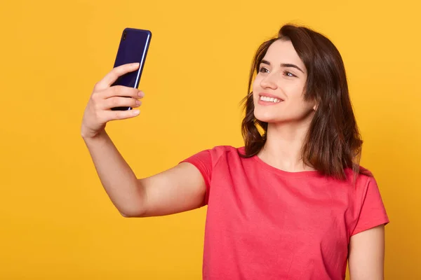 Horizontal shot of pretty girl taking self portrait with her smart phone, adorable lady looking devie's at camera with charming smile, attractive model posing isolated over yellow studio background. — Stock Photo, Image