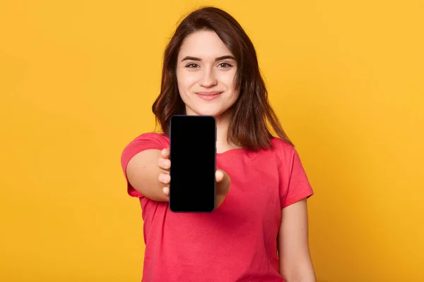 Tutup potret wanita muda yang menunjukkan tampilan ponsel dengan layar kosong hitam dan terlihat tersenyum langsung ke kamera yang terisolasi di atas latar belakang kuning. Salin ruang untuk advertismen . — Stok Foto