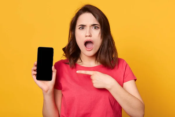 Portrait of good looking young brunette female showing smartphone screen and points at it with her finger, woman has schocked facial expression, posing with opened mouth. Copy space foer advertisment. — Stockfoto