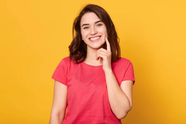 Jovem mulher caucasiana alegre com sorriso encantador suave, vestindo camisa vermelha, tendo aparência agradável, mantém o dedo indicador na bochecha, espera kiiss, sorri feliz, posa contra fundo amarelo . — Fotografia de Stock