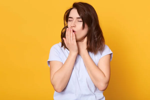 Portrait of tired girl with dark hair, standing and yawning with closed eyes, wearing casually clothing, posing isoalted over yellow studio background, wants to stay athome and lying in bed. — 스톡 사진