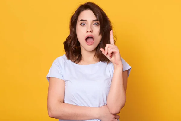 Retrato de mujer joven con cabello oscuro con camisa blanca casual de pie con la boca abierta, apuntando con el dedo hacia arriba para enfatizar la idea brillante, mujer attarctiva posando isoaled sobre fondo de estudio amarillo — Foto de Stock