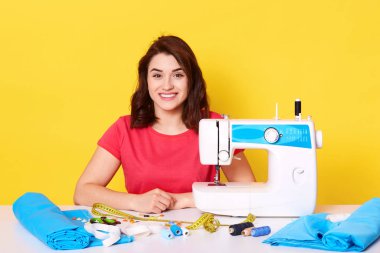 Çekici beyaz, çalışkan terzi stüdyosunda oturuyor, dikiş dikiyor, kırmızı günlük tişört giyiyor, masaya poz veriyor, doğrudan kameraya bakıyor, sarı arka planda..