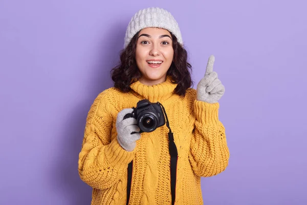 Foto horisontal gadis fotografer di atas dinding ungu menunjukkan dan menunjuk jari ke atas, memiliki ide bagus, melihat langsung ke kamera dengan mulut terbuka, mengenakan sweater kuning hangat, topi dan sarung tangan . — Stok Foto
