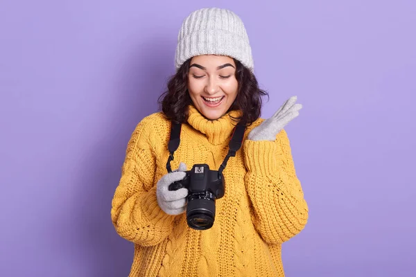 Charming girl watching photo in camera, posing over lilac background, female wearing warm knitted yellow sweater, gloves and cap, making photos, looks happy, keeping mouth open. Hobby concept. — 스톡 사진