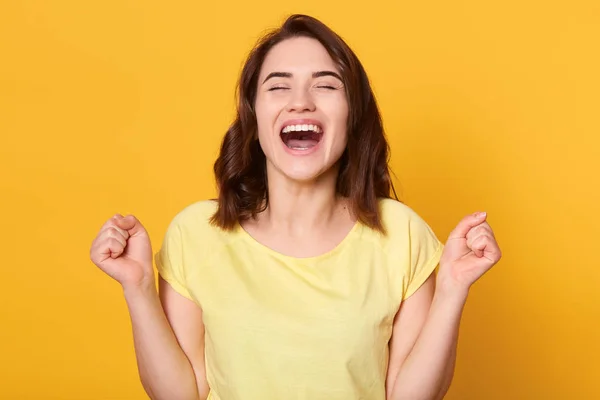 Close up portraiot of happy young woman with beautiful dark hair, clenching fist while looking directly at camera, wearing casual t shirt, isolated over yellow background. People emotions concept. — 스톡 사진
