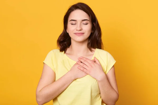 Image of satisfied generous brunette woman with charming smile, keeps both palms on chest, wearing casual yellow t shirt, standing against studio wall, being kind hearted. People, gratitude concept. — 스톡 사진