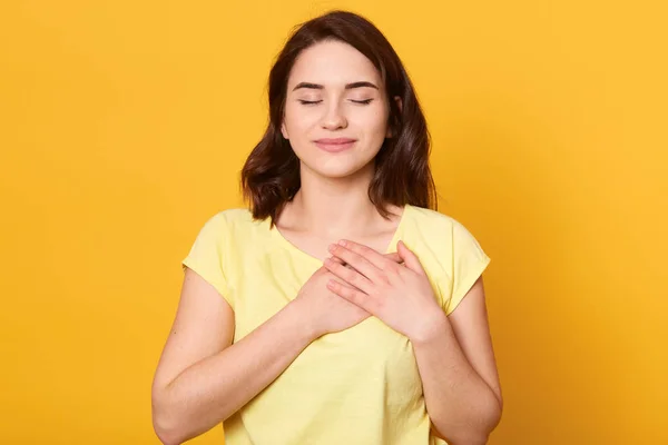 Imagen de mujer morena generosa satisfecha con una sonrisa encantadora, mantiene ambas palmas en el pecho, vistiendo una camiseta amarilla casual, de pie contra la pared del estudio, siendo amable de corazón. Gente, concepto de gratitud . —  Fotos de Stock