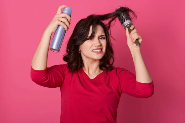 Close-up portret van ontevreden vrouwelijke kammen haar, woedend, maakt kapsel met haarborstel en spray, het dragen van rode shirt, ziet er boos, poseren geïsoleerd over roze achtergrond. Kapsel concept. — Stockfoto