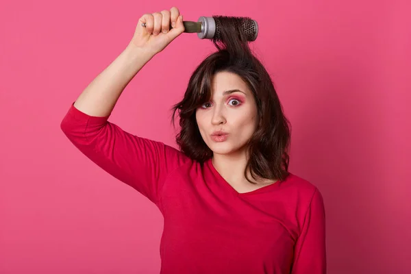 Fotografía horizontal de morena tensa sosteniendo pelo rebelde en peine, posando aislada sobre fondo de estudio rosa, manteniendo los labios redondeados, mirando a la cámara, usando una camisa roja casual. Concepto femenino . — Foto de Stock