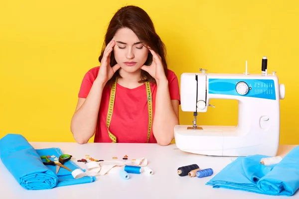 Horizontal shot of seamstress having pain in her head while working at workshop, young adorable dressmaker having headache, holding fingers on temples, surrounded sewing machine, piece of cloth. Stock Photo