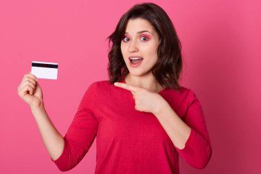 Picture of emotional good looking pretty female pointing with forefinger at credit card, having surprised facial expression, opening mouth, doing shopping online. Women and purchases concept. clipart