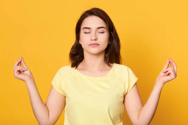 Image of peaceful good looking brunette wearing yellow t shirt, raising hands, having meditation, closing eyes, being alone, keeping silence, sitting isolated over yellow background in studio. — 스톡 사진