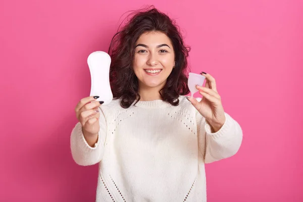 Retrato de estudio interior de mujer rizada linda magnética alegre con copa menstrual y almohadilla de higiene en ambas manos, mirando directamente a la cámara, usando suéter blanco. Concepto de menstruación . —  Fotos de Stock