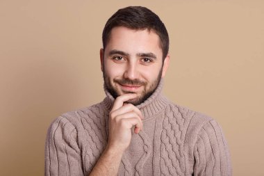 Bej duvara karşı poz veren mutlu genç adamın portresi. Sıcak kış süveteri giyen neşeli bir adam, kameraya doğrudan bakan sakallı bir adam, çenesinin altında parmağı olan sakallı adam, kendinden emin görünüyor.