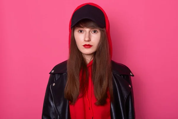 Horizontal shot of young beautiful fashionable teenager girl wearing leather jacket, red hoody and black cap, looking directly at camera, looks confident and serious, model posing with red lips.
