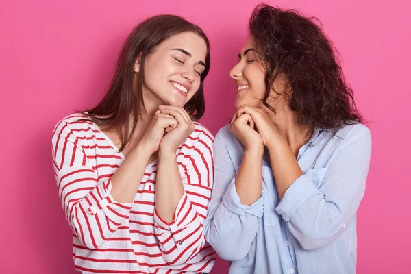 Horizontal indoor picture of two cute smiling peaceful good looking friends, closing eyes, clenching hands, enjoying rest, spending leisure time together, standing isolated over pink background. — 스톡 사진