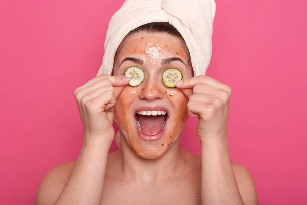 Studio shot de mujer satisfecha se aplica rodajas de pepino en los ojos y tiene máscara de arañazo en la cara, tiene expresión feliz, abre la boca con amplitud, haciendo procedimientos de belleza en casa, usa toalla blanca . — Foto de Stock