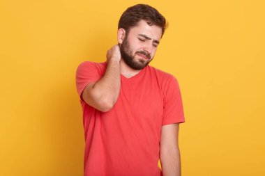 Çekici, sakallı, boynu ağrıyan, kırmızı tişört giyen, boynuna dokunan ve sarı arka planda durarak olumsuzluğunu ifade eden genç bir adam..