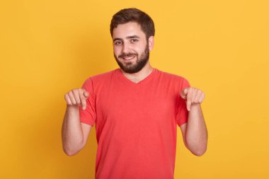 Doğrudan kameraya bakan, parlak kırmızı tişörtlü, beyaz stüdyo duvarına yaslanmış, ön parmaklarını aşağı gösteren genç bir erkeğin yatay görüntüsü. Reklam için alanı kopyala.