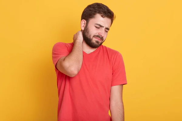 Bild eines attraktiven bärtigen Mannes, der Nackenschmerzen verspürt, frustrierter junger Mann, der ein rotes lässiges T-Shirt trägt, seinen Hals berührt und vor gelbem Studiohintergrund Negativität ausdrückt. — Stockfoto