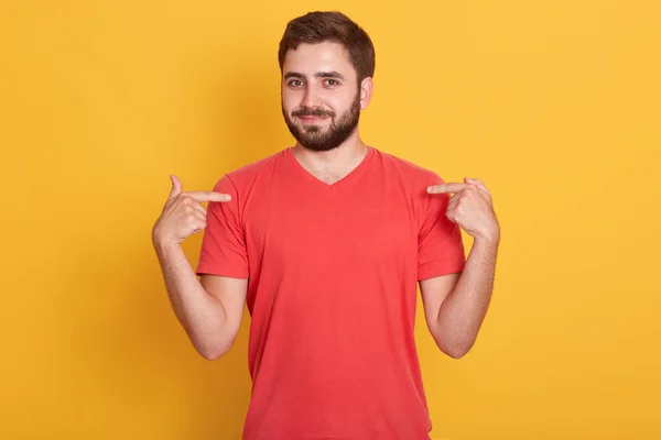 Πορτρέτο του όμορφος γενειοφόρος νεαρός άνδρας δείχνει τον εαυτό του, ελκυστική ευρωπαϊκή αρσενικό στέκεται πάνω από κίτρινο φόντο, έχει ευχάριστη εμφάνιση, φορώντας κόκκινο casual t πουκάμισο. Αντιγραφή χώρου για διαφήμιση. — Φωτογραφία Αρχείου