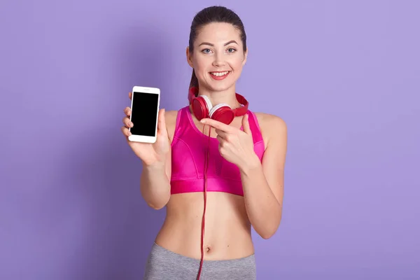 Image d'une femme sportive joyeuse et adorable faisant un geste, pointant du doigt un smartphone avec un écran vide, tenant son appareil d'une main, souriant sincèrement, étant de bonne humeur. Concept technologique . — Photo