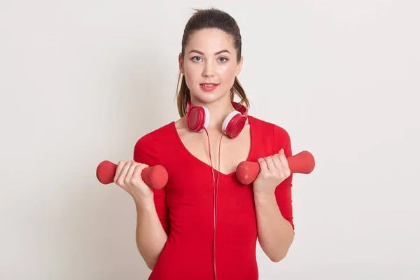 Horizontal indoor portrait of tired self-motivated brunette with ponytail looking directly at camera, having headphones around neck, holding red dumbbells, doing exercises in order to lose weight. — Stock Photo, Image