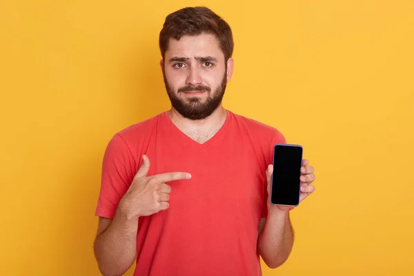 Joven enojado viste camiseta roja casual posando aislado sobre fondo amarillo en el estudio, chico guapo señalando con su dedo índice en el teléfono móvil con la pantalla vacía en blanco. . — Foto de Stock