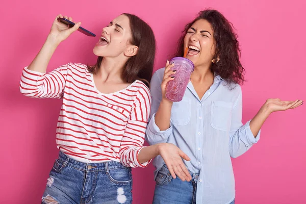 Foto close up gadis-gadis bahagia menari dan bersenang-senang sambil mendengarkan musik dan bernyanyi favorit, menggunakan telepon dan botol air instan mikrofon, terisolasi di atas latar belakang merah muda . — Stok Foto