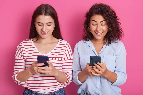 Horizontal shot of attractive Caucasian women standing isolated over pink background with phones in their hands, looking ar device's screens with happy axpressions, ladies wearing casual clothing. — Stock Photo, Image