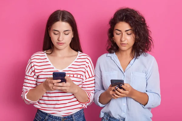 Primer plano retrato de dos niñas visten trajes casuales, de pie aislado sobre fondo rosa pastel. Dos mujeres usando teléfonos móviles, escribiendo mensajes SMS, pareciendo concentradas. Concepto tecnológico . — Foto de Stock