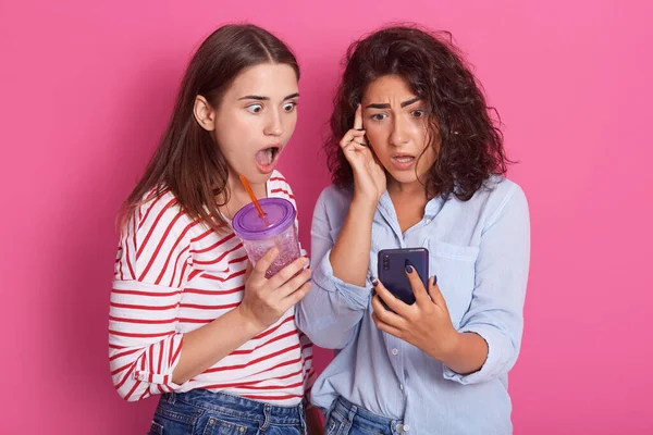Image of two shocked young ladies posing over rosy studio background. Looking at phone\'s screen with widely opened mouths and astonished facial expressions, check social network. Gossip concept.