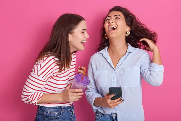 Horizontalshot de mujeres jóvenes bonitas riendo felizmente, tienen teléfono y botella de agua en las manos, con ropa elegante, posando aislado sobre fondo de estudio rosa. Alto espíritu, concepto de amistad . — Foto de Stock