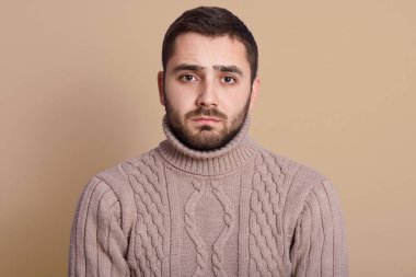 Stüdyoda bej duvara yaslanmış sakallı çekici bir gencin yatay görüntüsü. Yüzünde ciddi bir ifadeyle kameraya bakıyor. Örgü kazak giyiyor..