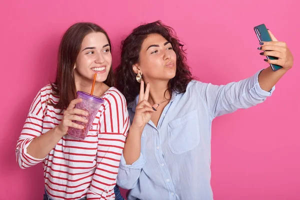 Horizontal shot of two girls friends taking selfie with smartphone, blowing kiss, shoving v sign, standing with water bottle, models posing isolated over rose background. Technology concept.