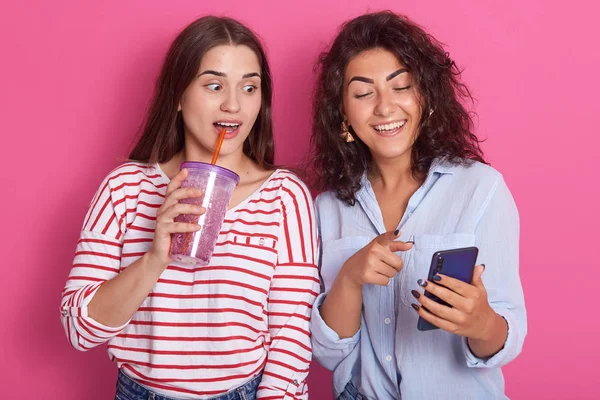 Imagen de dos mujeres jóvenes de buen aspecto de pie aisladas sobre el fondo del estudio rosa, una señora sosteniendo el teléfono celular y otra sostiene la botella de agua y bebidas, mirando la pantalla del dispositivo con interst . — Foto de Stock