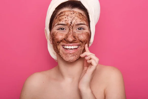 Indoor shot of happy smiling girl with white towel on her head, applying mask to her face, doing beuity procedures at home in morning, standing wirh bare shoulders isolated over rosy background. — Stock Photo, Image