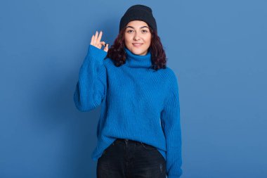 Kapalı alanda hoş bir esmerin olumlu bir jest yaptığını gösteren bir resim. Doğrudan kameraya bakıyor, koyu mavi kazak ve şapka giyiyor, pantolon giyiyor. İnsanlar ve vücut dili kavramı.