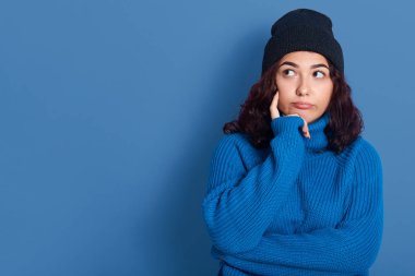 Düşünceli, tapılası, güzel bir kadının yan yan bakan, parmaklarıyla Cheeck 'e dokunan, üzgün, düşünceli bir yüz ifadesi olan kapalı mekanda çekilmiş resmi. Reklam için eşdeğer alan.