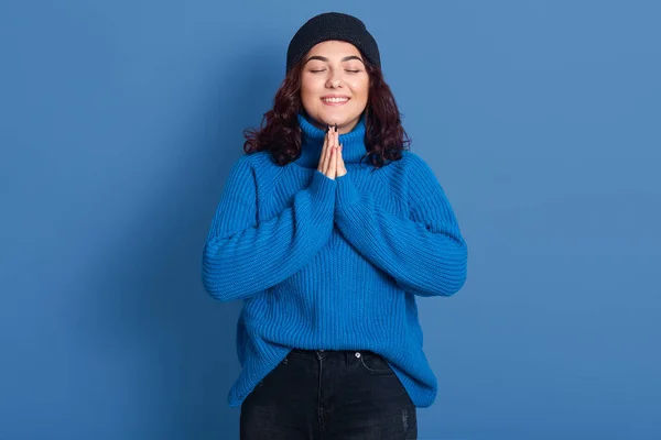 Bild av glada bedårande ung dam står med slutna ögon isolerade över blå bakgrund i studio, bär mörkblå tröja och hatt, svarta byxor, ler uppriktigt. Känslor koncept. — Stockfoto