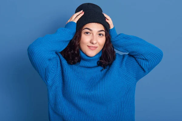 Imagen horizontal de una hermosa mujer guapa vestida de azul oscuro y suéter, mirando directamente a la cámara, poniendo las manos en la cabeza, con el pelo rizado negro, estando de buen humor. Concepto de invierno . — Foto de Stock