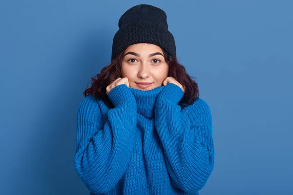 Retrato de primer plano de una joven carismática magnética de buen aspecto mirando directamente a la cámara, tocando el suéter azul con las manos, con expresión facial pacífica, usando suéter azul y sombrero . — Foto de Stock