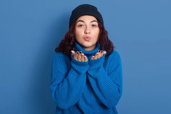 Image of energetic active lovely lady sending kisses, raising hands, being sincere, wearing dark blue sweater and hat, standing isolated over blue background in studio. People and emotions concept. — ストック写真