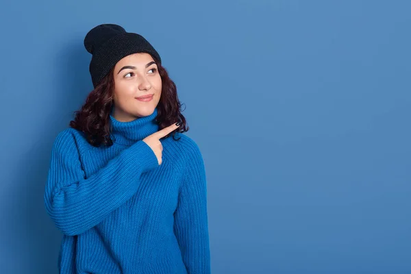 Indoor image of charismatic lovely positive attractive female looking aside, making gesture, pointing with forefinger sideways, wearing dark blue hat and sweater. Copyspace for advertisement. — 스톡 사진