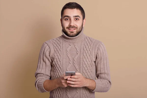 Image of pleasant positive young man with beard and black hair smiling sincerely, holding smartphone in both hands, looking directly at camera, having peaceful facial expression. Emotions concept. — Stock Photo, Image