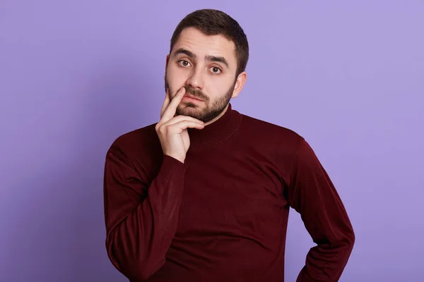 Horizontal shot of thoughtful focused sweet bearded man looking directly at camera, standing isolated over lilac background in studio, wearing dark red sweatshirt. People and emotions concept. — Stock Photo, Image